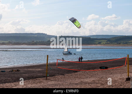 Un kite surfer sur un jour de vent à l'estuaire de la rivière Exe avec les marcheurs de la plage, un voilier et un filet de volley-ball dans l'avant-plan. Banque D'Images