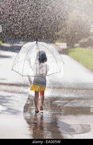 Vue arrière de barefoot girl exerçant son parapluie marche à travers street flaque Banque D'Images