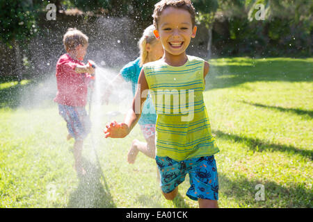 Trois enfants courir après eux dans jardin avec water sprinkler Banque D'Images