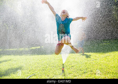 Girl jumping over water sprinkler dans jardin Banque D'Images