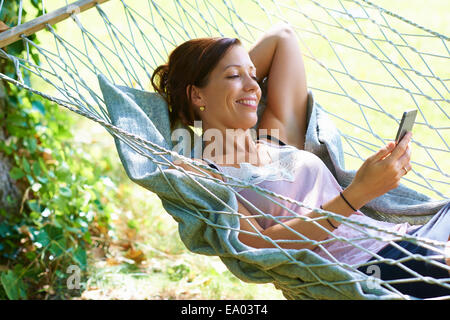 Mid adult woman reclining dans jardin Hamac looking at smartphone Banque D'Images