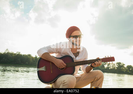 Jeune homme assis au bord du lac à jouer de la guitare Banque D'Images