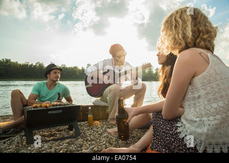 Jeune homme assis au bord du lac avec des amis jouer de la guitare Banque D'Images