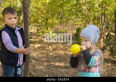 Jolie petite fille blonde sauter un ballon jaune coloré partie à l'extérieur dans le jardin vu par un jeune garçon tenant un t Banque D'Images