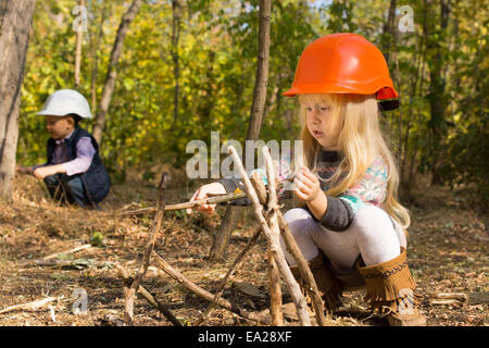 Cute little girl la construction d'un tipi en bois s'accroupissant sur le sol dans les bois dans son casque tandis qu'un jeune garçon travaille à l'arrière Banque D'Images