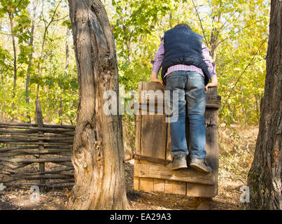 Petit Garçon jouant sur une ancienne porte rustique en bois entre deux troncs d'arbre en équilibre sur elle avec son dos à la caméra face à Banque D'Images