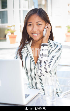 Young Asian woman dans la police d'un ordinateur portable et parlant au téléphone Banque D'Images