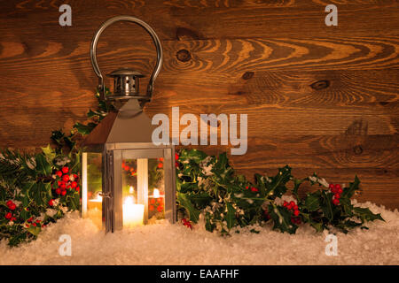 Lanterne à bougie de Noël dans la neige entouré par Holly avec fruits rouges contre un arrière-plan en bois rustique. Banque D'Images