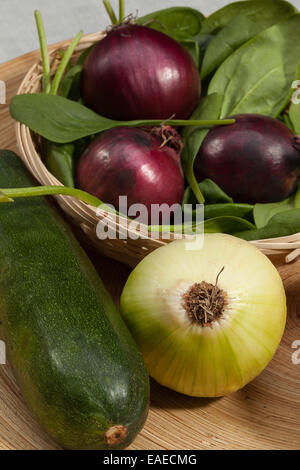 Un mélange de fruits et légumes d'automne : oignons rouges, oignon blanc, courgette dans un panier en osier sur fond boisé. Banque D'Images