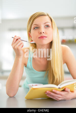Young woman eating yoghurt en cuisine et reading book Banque D'Images