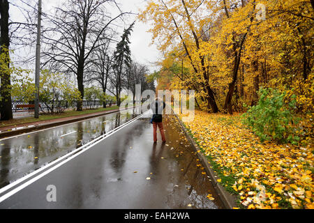 Fille qui marche sur la route mouillée parmi une forêt d'automne couverte de feuilles jaunes Banque D'Images