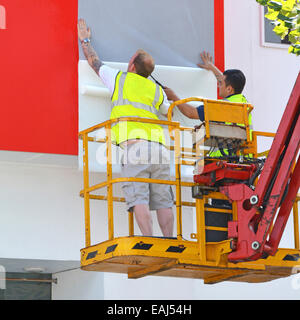 Grue de la plate-forme d'accès en cours d'utilisation par des ouvriers feuilles de fixation formant graphique une grande annonce sur le côté du bâtiment d'affichage Banque D'Images