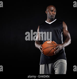 Portrait de joueur de basket-ball noir avec ballon à l'extérieur contre fond noir. Jeune homme fort avec le basket-ball. Banque D'Images