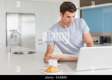 L'homme avec du papier journal à l'aide d'ordinateur portable au comptoir de la cuisine, tasse de café et un croissant en premier plan Banque D'Images