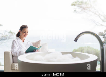 Smiling woman in bathrobe reading book in bathroom Banque D'Images