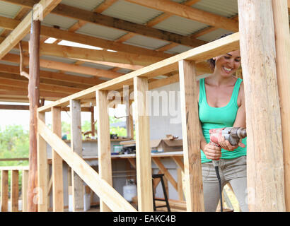 Smiling woman drilling in plank in house under construction Banque D'Images