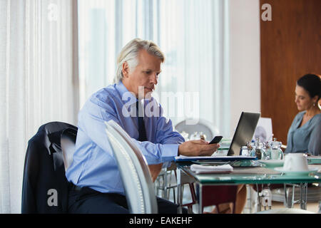 L'homme d'affaires à maturité à l'aide de téléphone mobile au restaurant table, woman in background Banque D'Images