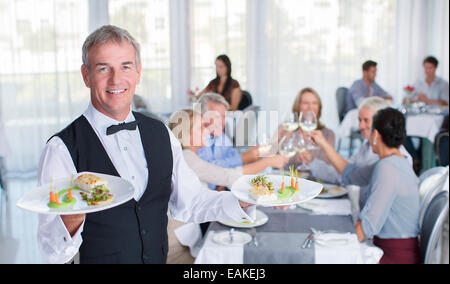 Portrait of waiter holding plaque avec fantaisie les repas, personnes à tables de restaurant en arrière-plan Banque D'Images