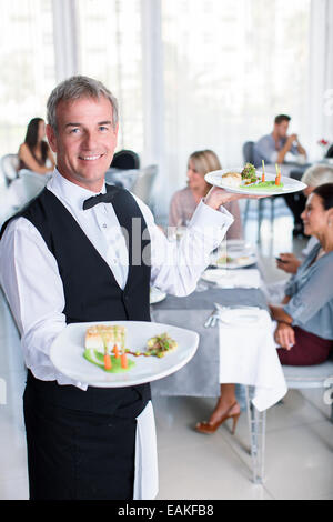 Portrait of smiling waiter holding plats fantaisie, les gens assis à des tables de restaurant en arrière-plan Banque D'Images