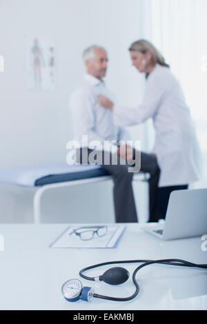 Jauge de pression artérielle, ordinateur portable, fichier et lunettes sur 24 dans le cabinet du médecin, female doctor examining patient in background Banque D'Images