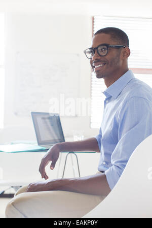 Portrait of young smiling man wearing glasses and blue shirt sitting at desk with laptop Banque D'Images