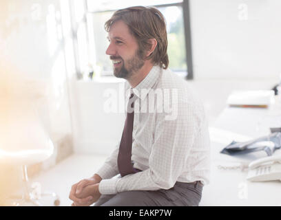 Smiling businessman wearing chemise et cravate sitting on desk in office Banque D'Images