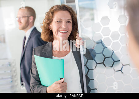 Portrait of woman holding documents debout à la porte de verre, d'un collègue en arrière-plan Banque D'Images