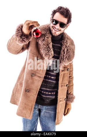 Un joyeux jeune homme portant un manteau de fourrure de mouton cher isolé sur un fond blanc et tenant une boule de noël rouge Banque D'Images