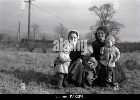 Années 1950, tableau historique, posant pour une photo à l'extérieur à genoux sur l'herbe dans un champ, deux mères happy smiling holding leurs trois petits enfants, England, UK. Banque D'Images