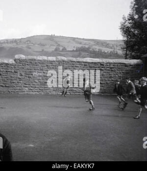 Années 1950, photo historique d'un groupe de garçons d'école jouant au football avec un petit ballon dans le terrain de jeu contre un mur de pierre dans une école rurale, Angleterre, Royaume-Uni. Banque D'Images