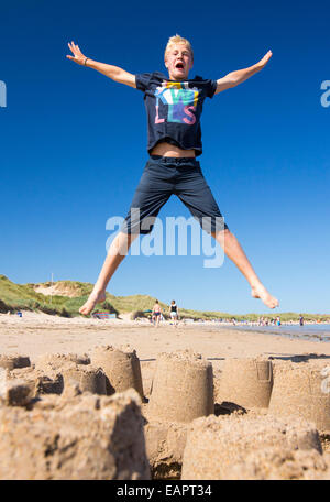 Un garçon sautant au dessus d'un château de sable sur la plage de Bamburgh, Northumberland, Angleterre. Banque D'Images