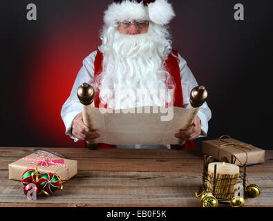 Santa Claus holding sa coquine et belle liste sur un rouleau de papier sulfurisé. Santa est assis à une table en bois avec des présents et Banque D'Images