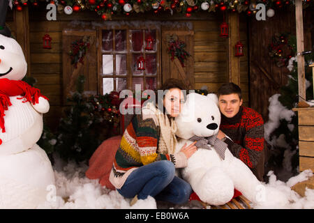 Jeune couple assis dans des tenues d'hiver Hugging blanc hiver Porter Poupée à Maison en bois décorée avec de grandes Snowman et divers Chr Banque D'Images