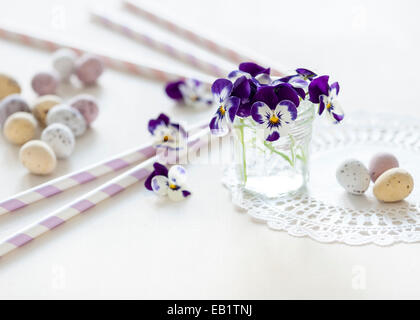 Petit bouquet de pensées dans un bocal en verre sur un napperon sur le tableau blanc, avec des mini-oeufs et pailles pastel tous baigné de soleil Banque D'Images