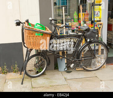 Vieux vélo noir avec épicerie panier en osier & enseigne publicitaire intégrée à l'extérieur du châssis boutique matériel à Brecon, Wales Banque D'Images