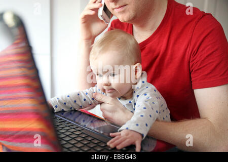 Un père travaille sur son ordinateur portable à partir de la maison tout en tenant son bébé de huit mois. Banque D'Images