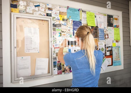 Young woman photographing tableau d'affichage communautaire avec smartphone Banque D'Images