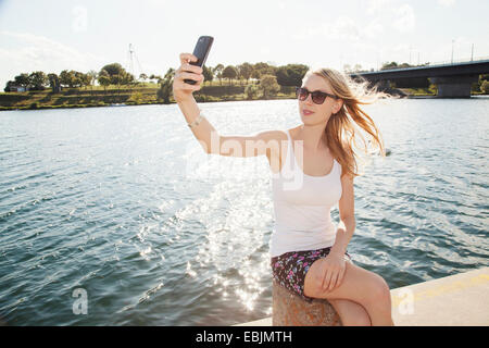 Jeune femme assise à la Riverside en tenant selfies smartphone, l'île du Danube, Vienne, Autriche Banque D'Images