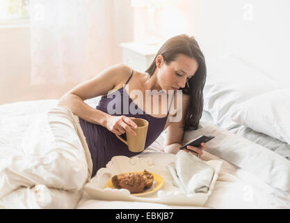 Woman having breakfast in bed, using mobile phone Banque D'Images