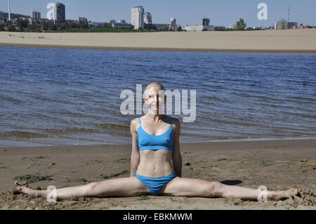 Bald woman sitting in split, ou conasana upavistha, sur une plage de sable, sur les toits de la ville dans la distance Banque D'Images