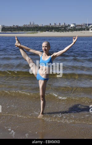 Bald woman standing in yoga position appelée-Big-Toe Hand-To prolongée Pose ou utthita hasta padangusthasana sur une plage de sable fin Banque D'Images