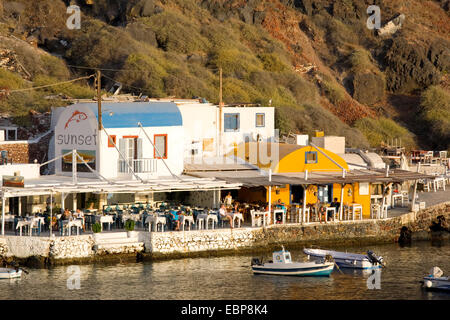 Ia, Santorin, Grèce, sud de la Mer Egée. Tavernes au bord de l'eau à la baie Ammoudi éclairées par le soleil couchant. Banque D'Images