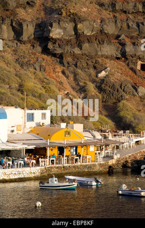 Ia, Santorin, Grèce, sud de la Mer Egée. Tavernes au bord de l'eau à la baie Ammoudi éclairées par le soleil couchant. Banque D'Images