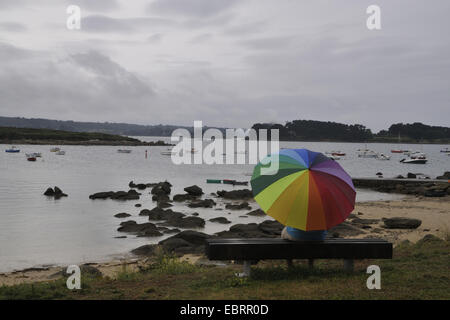 Homme assis sous la pluie avec un parapluie coloré sur un banc et regarda la mer, maison de vacances pluvieux, France, Bretagne Banque D'Images