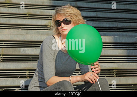 Jeune femme blonde avec des lunettes et green balloon assis sur les escaliers, Allemagne Banque D'Images