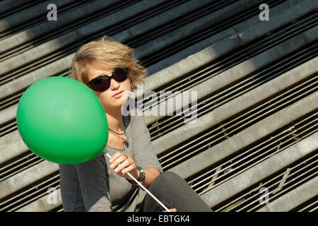 Jeune femme blonde avec des lunettes et green balloon assis sur les escaliers, Allemagne Banque D'Images