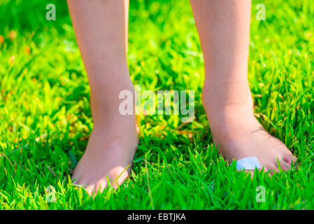 Enfant pieds nus avec un plâtre sur un pied se dresse sur l'herbe luxuriante Banque D'Images