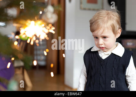 Petit garçon perdu dans ses pensées alors qu'il se tenait devant un arbre de Noël avec un burning sparkler à c Banque D'Images