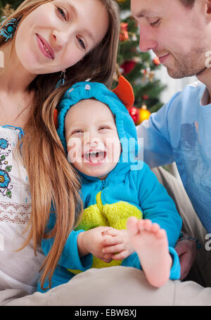 Les jeunes parents couvrir avec l'appareil photo tout en embrassant la main de bébé, fils habillés en costume de monstre peu rire, le temps de Noël. Banque D'Images