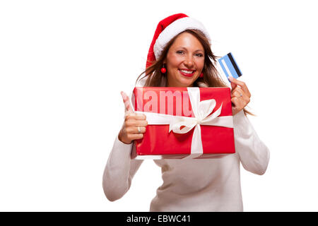 Smiling woman wearing a Santa hat rouge l'achat de cadeaux de Noël sur une carte bancaire brandissant une giftwrapped fort rouge avec un sourire heureux et un coup de geste de succès, isolated on white Banque D'Images